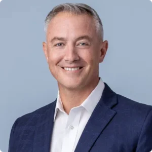 Man in navy suit jacket, white button down, smiling at camera.