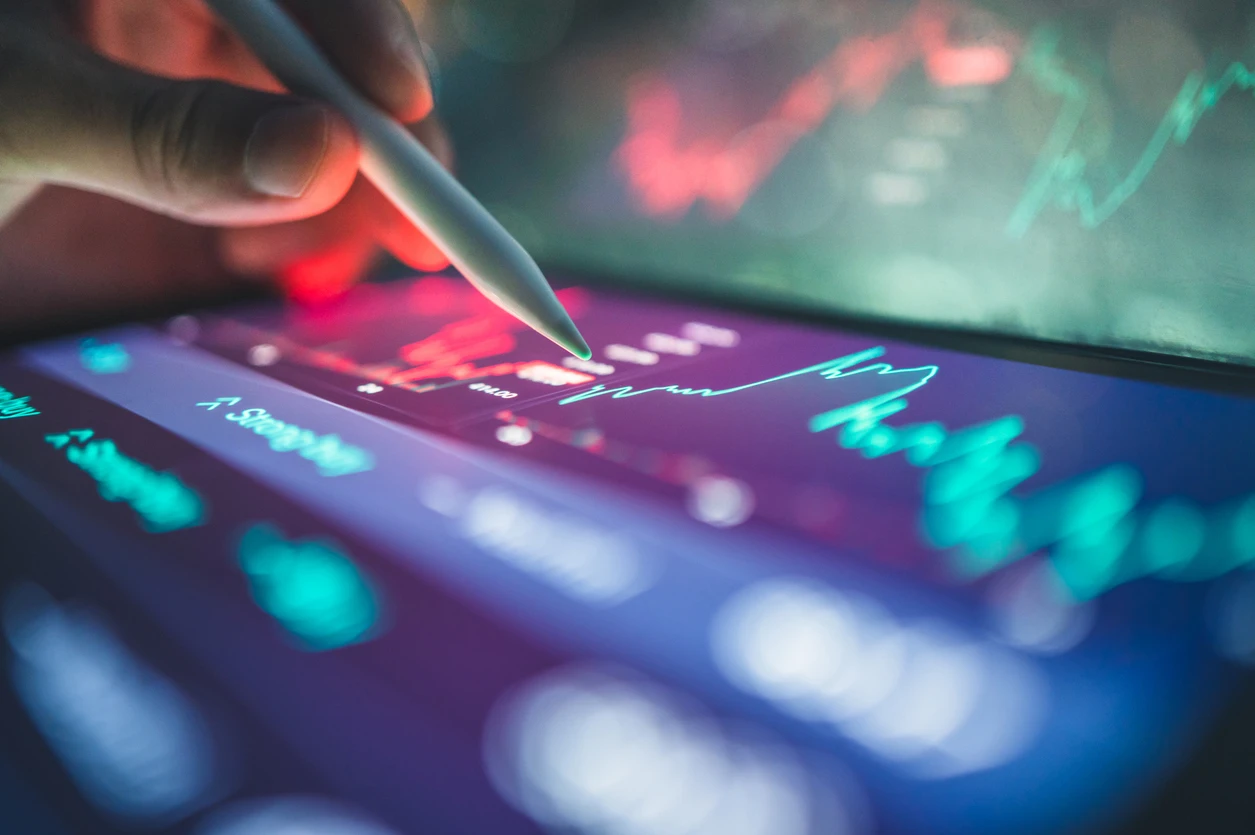 close up of a hand holding a white stylus writing on a tablet. The display glows with dark purples, teal and blue and displays financial charts and figures.